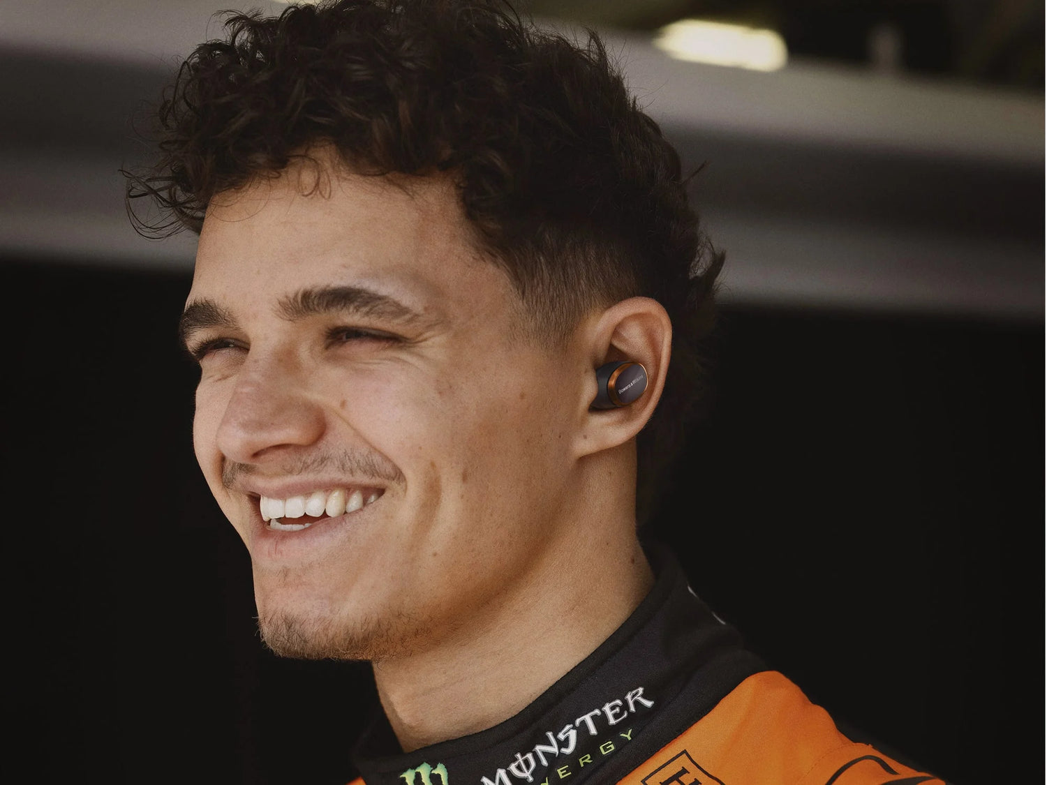 Person wearing a racing suit with a visible brand logo, smiling, wearing Bowers & Wilkins PI8, McLaren Edition headphones.