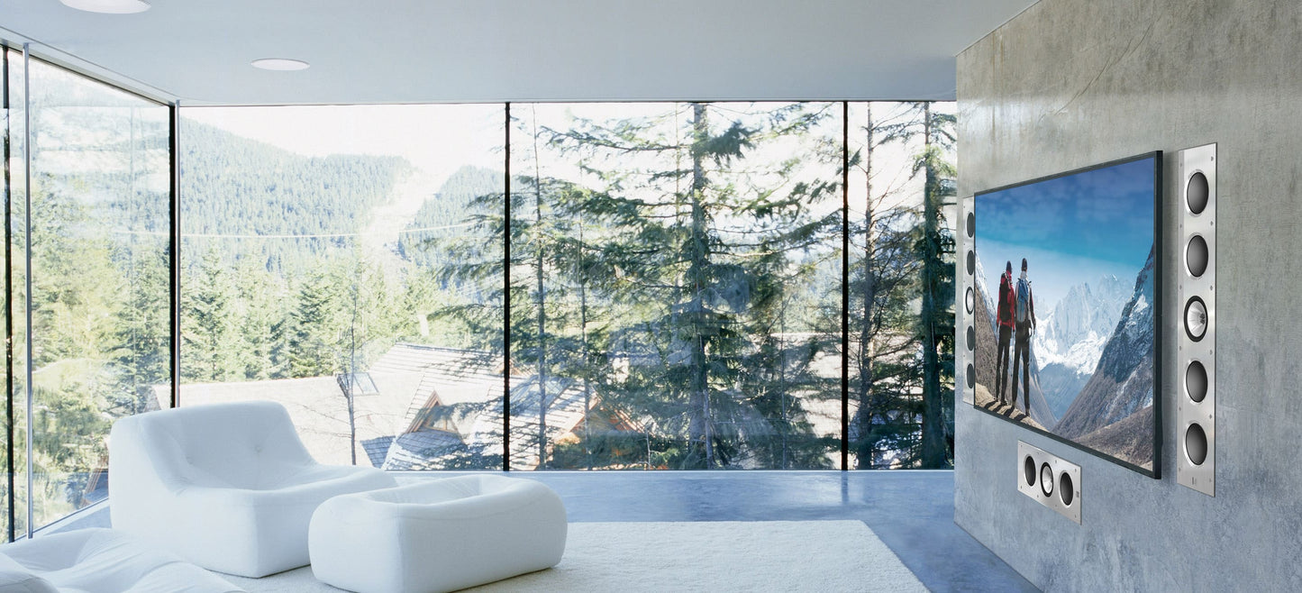 Modern living room with a large TV featuring KEF Ci200RR-THX Extreme In-Ceiling displaying a mountain scene, surrounded by large windows with a forest view.