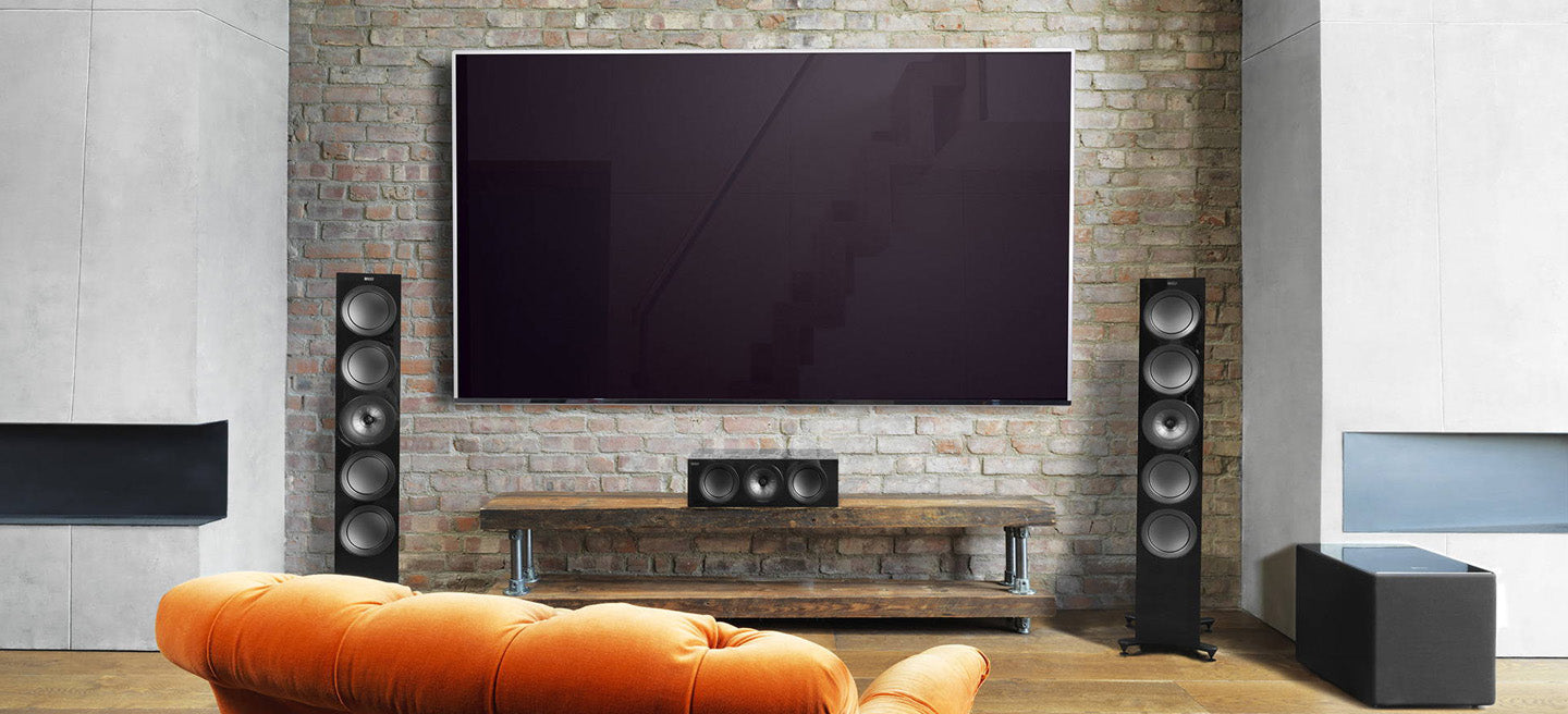 Modern living room with a large flat-screen TV on a brick wall, orange sofa, and speakers and KEF 8b referene subwoofer.