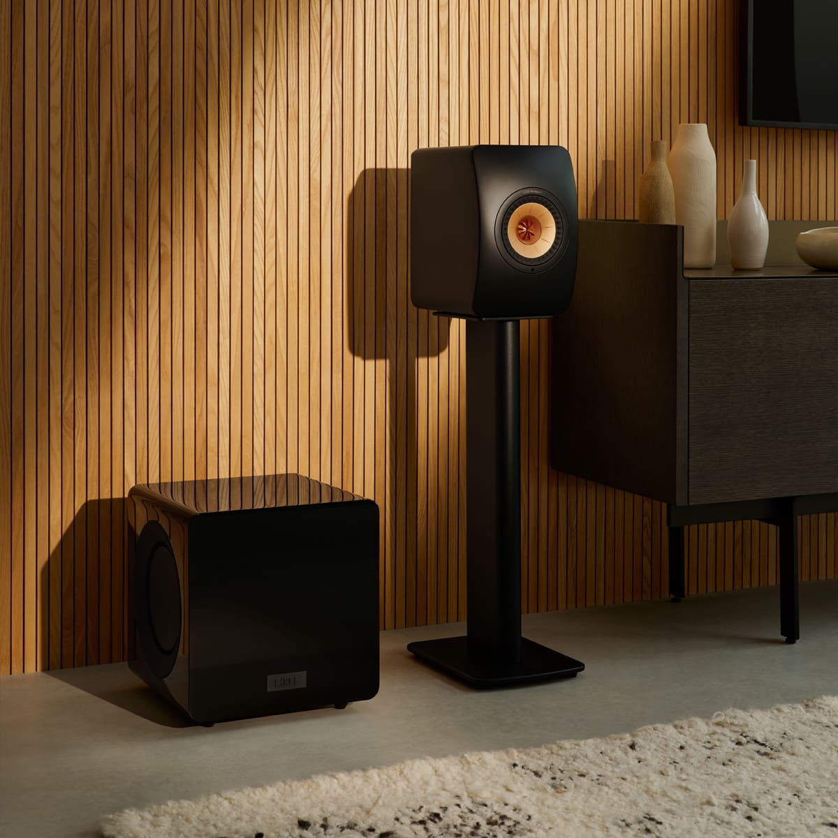 Black speaker and a black KEF KC92 Dual 9” Subwoofer on stands against a wooden wall with decorative vases.