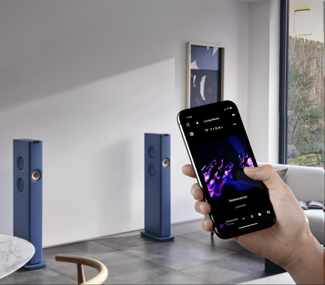 Person holding a smartphone with music app on screen in a modern living room with KEF LS60 Wireless Speakers.