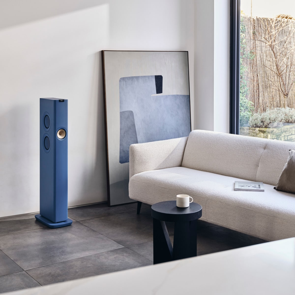 Modern living room with a blue floor-standing KEF LS60 Wireless Speakers, white sofa, and abstract art.