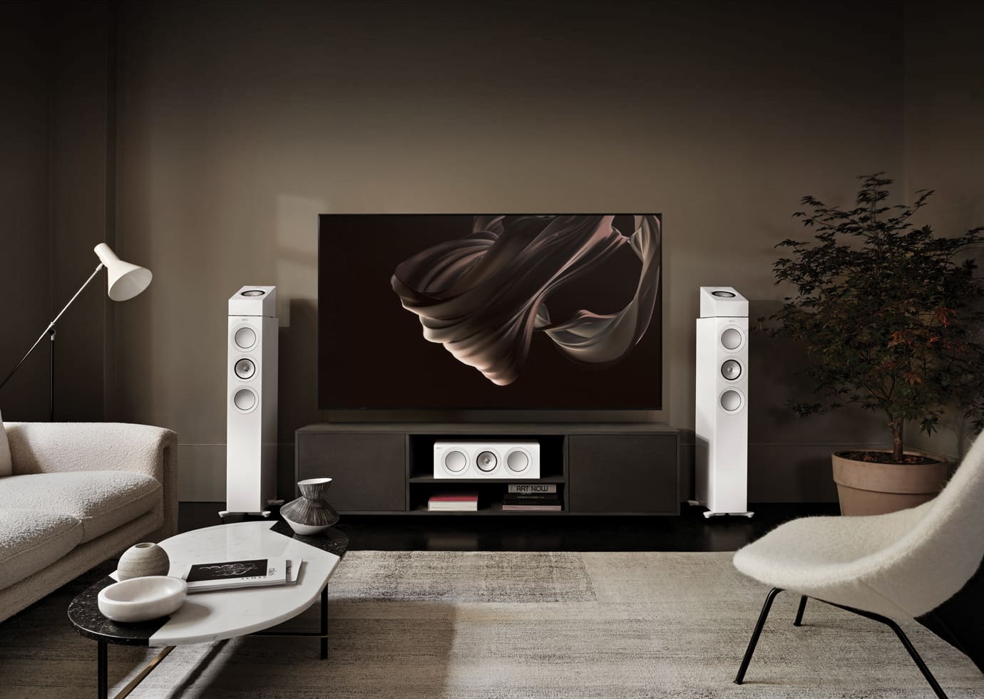 Modern living room with a large TV on a stand, flanked by KEF R7 Meta Floor Standing Speaker, in a dimly lit room.