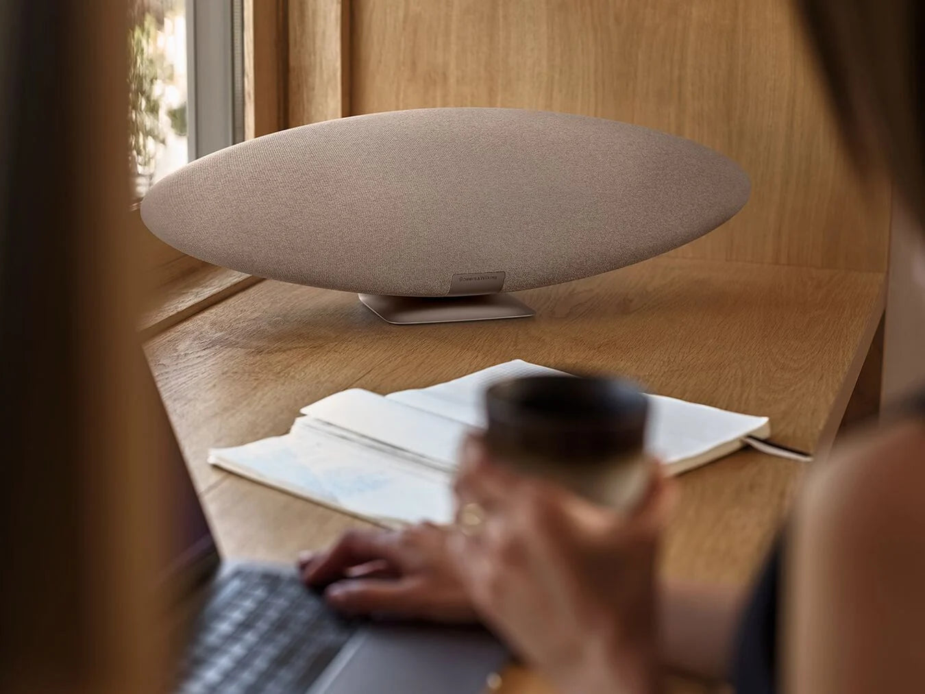 Person using a laptop with a Bowers & Wilkins Zeppelin Pro Wireless Speaker in solar gold on a wooden table.