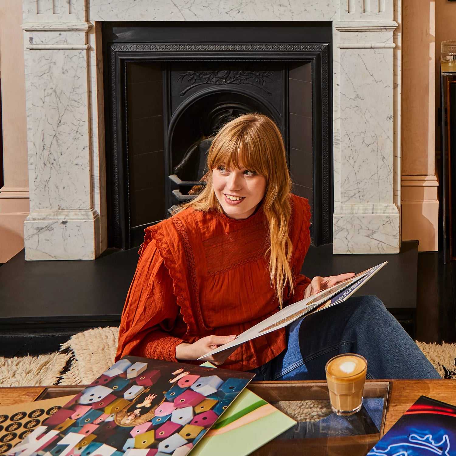 Woman sitting by a fireplace holding a vinyl, with a coffee cup on a table in front of her.