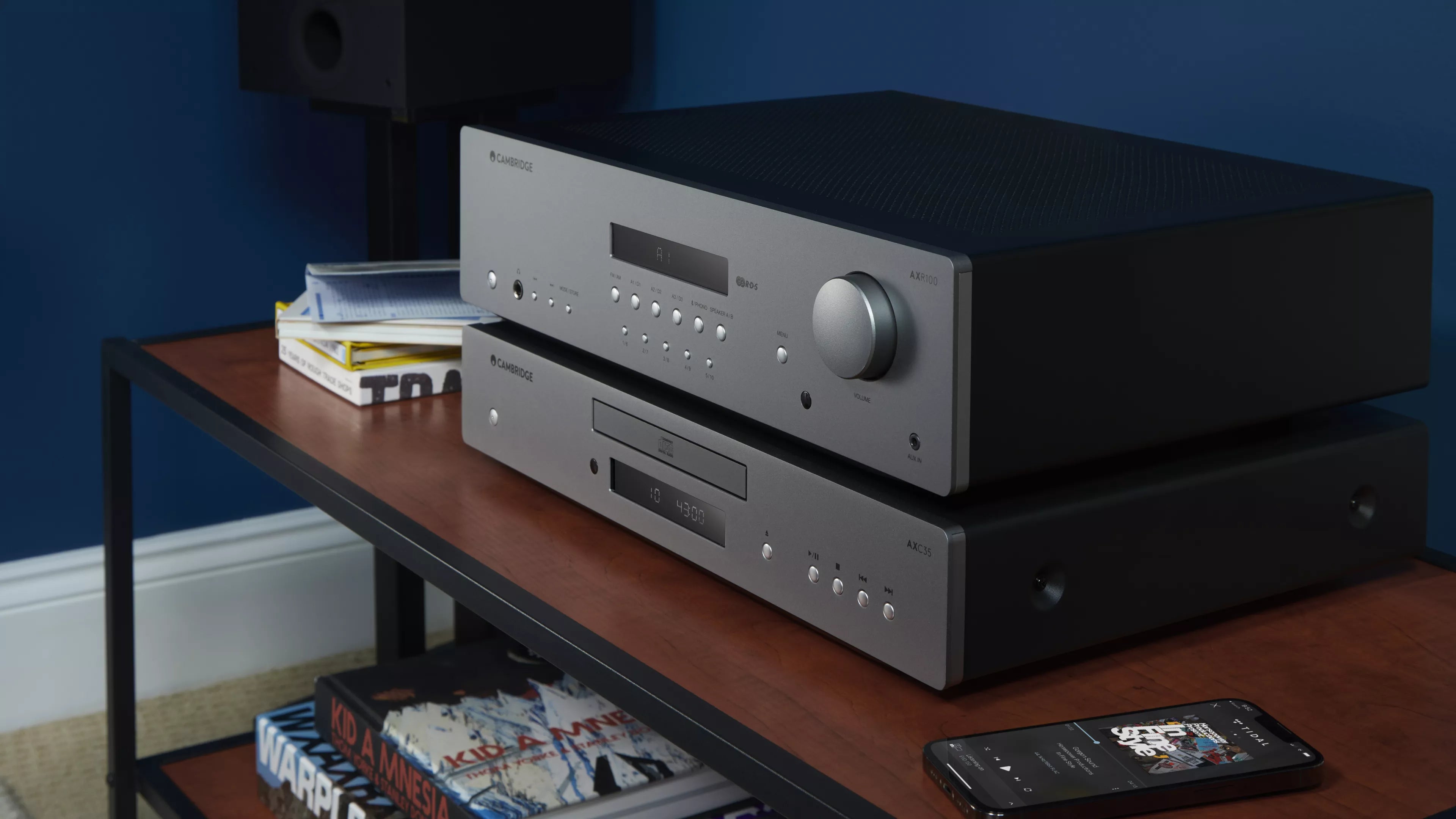 Audio equipment with a Cambridge Audio AXR100 Stereo Receiver on a wooden shelf against a blue wall.