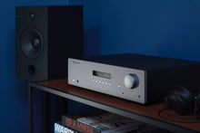 Audio equipment including a speaker, Cambridge Audio AXR100 Stereo Receiver, and headphones on a shelf against a blue wall.