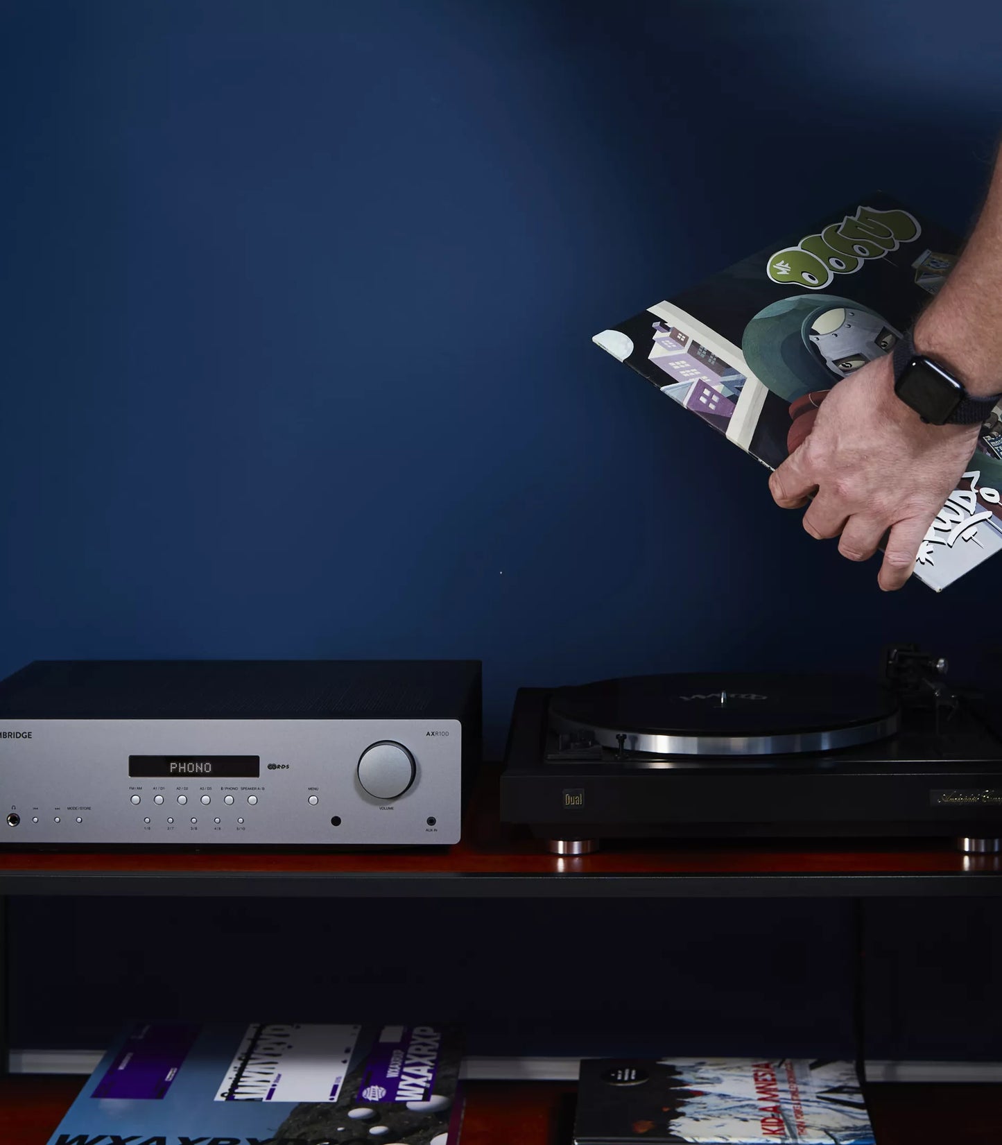 Person placing a vinyl record on a turntable with a dark blue wall in the background next to a Cambridge Audio AXR100 Stereo Receiver.