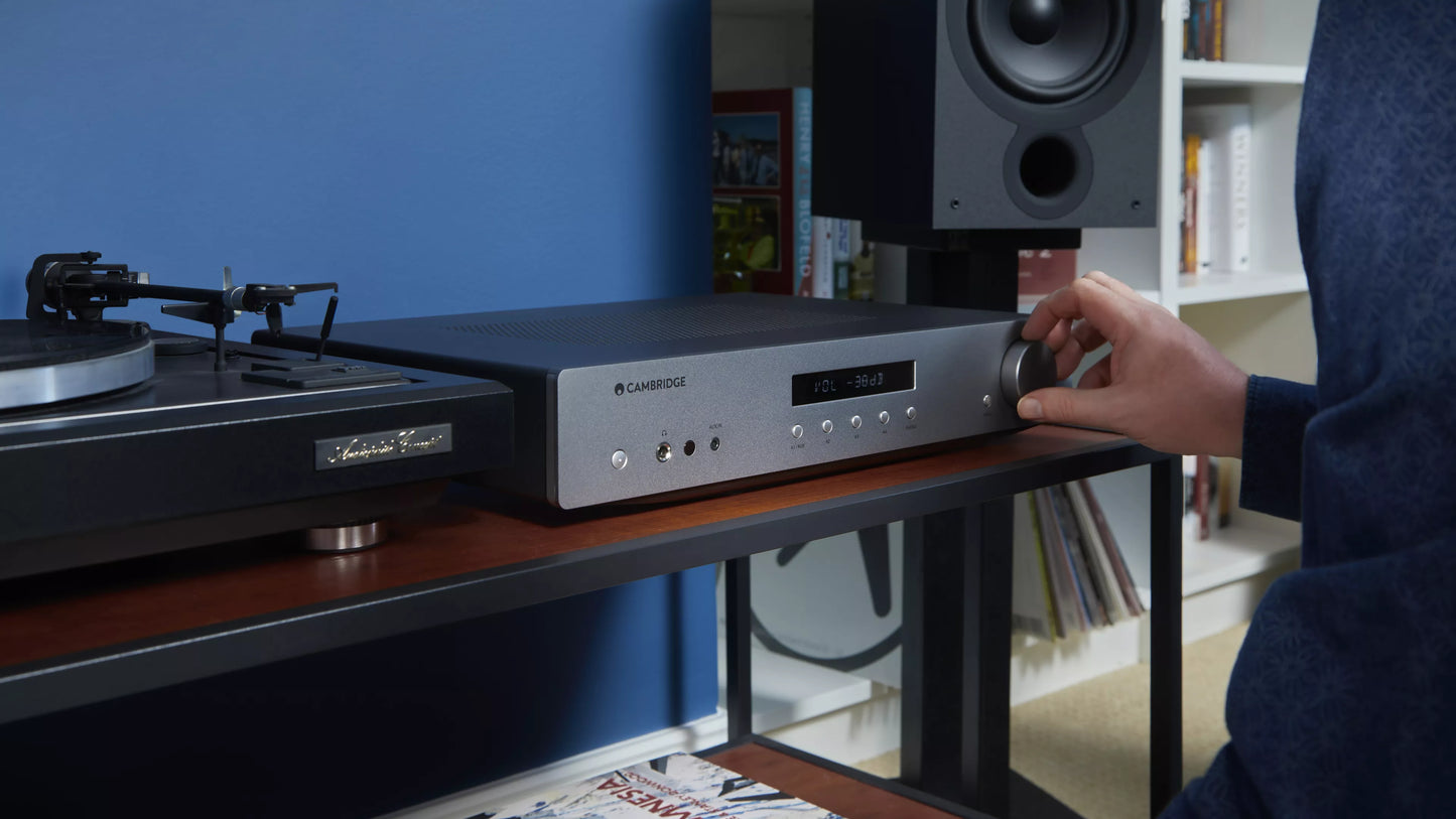 Person adjusting equipment on a turntable and Cambridge Audio AXA35 Integrated Amplifier with a blue wall and bookshelf in the background.
