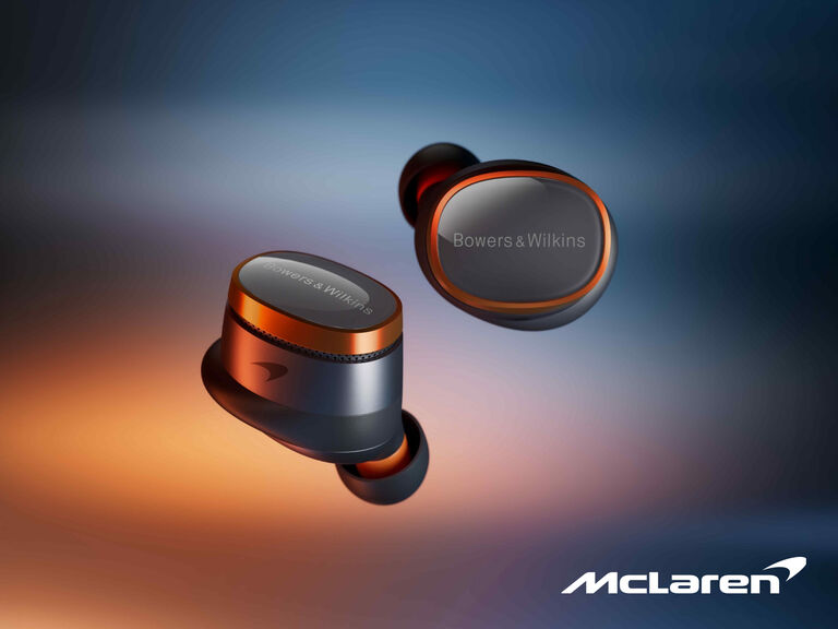 A pair of Bowers & Wilkins PI8 McLaren Edition in-ear headphones in Galvanic Grey with Papaya orange accents on a blurred colorful background.