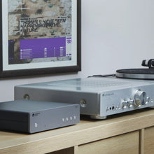 Cambridge Audio MXN10 Compact Network Player on a wooden surface with a framed poster in the background.