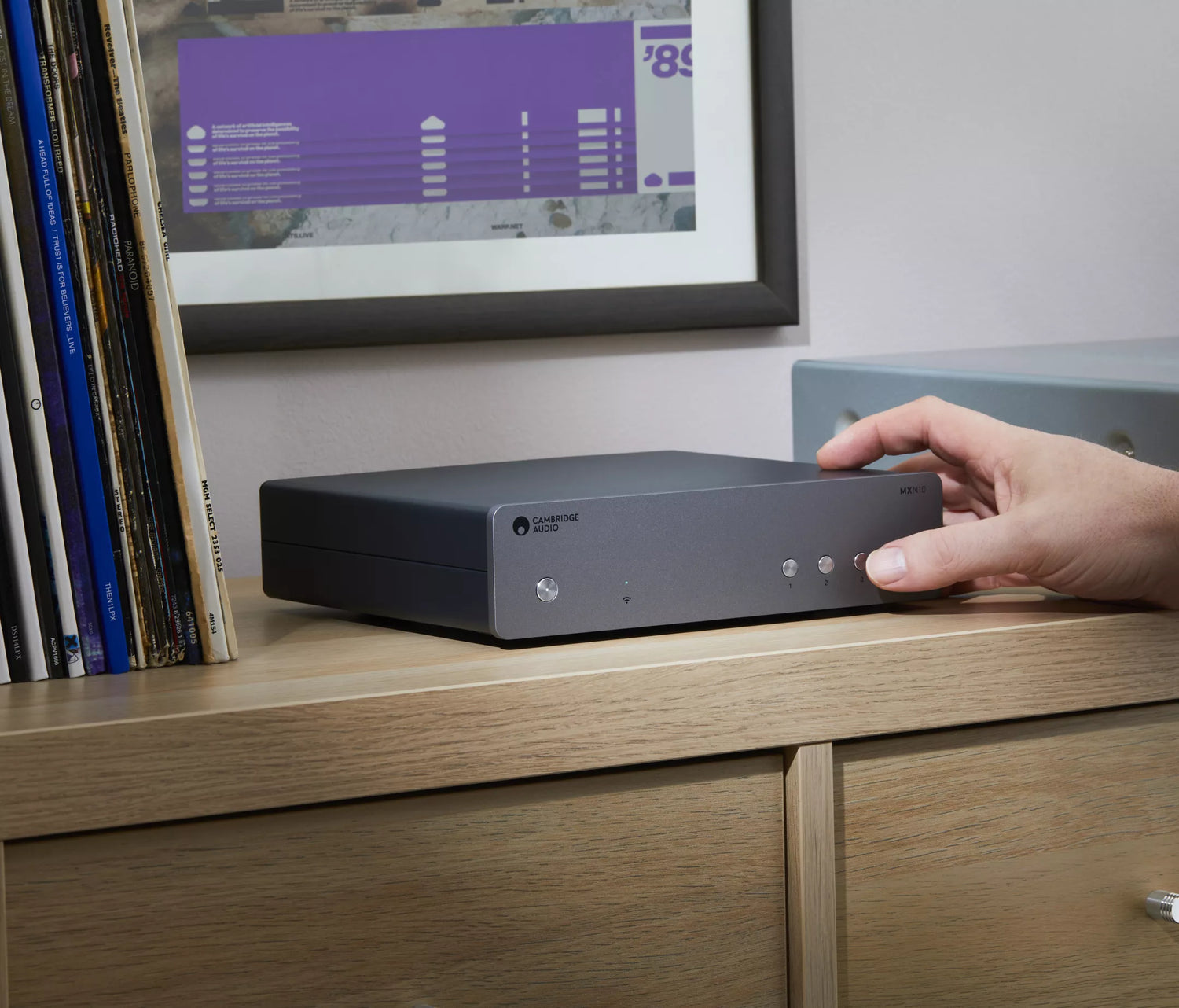Hand interacting with a Cambridge Audio MXN10 Compact Network Player on a wooden surface with a computer screen in the background.
