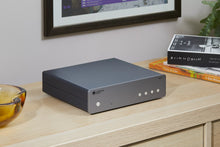 Gray Cambridge Audio MXN10 Compact Network Player on a wooden surface with books and a framed picture in the background in a modern living room.