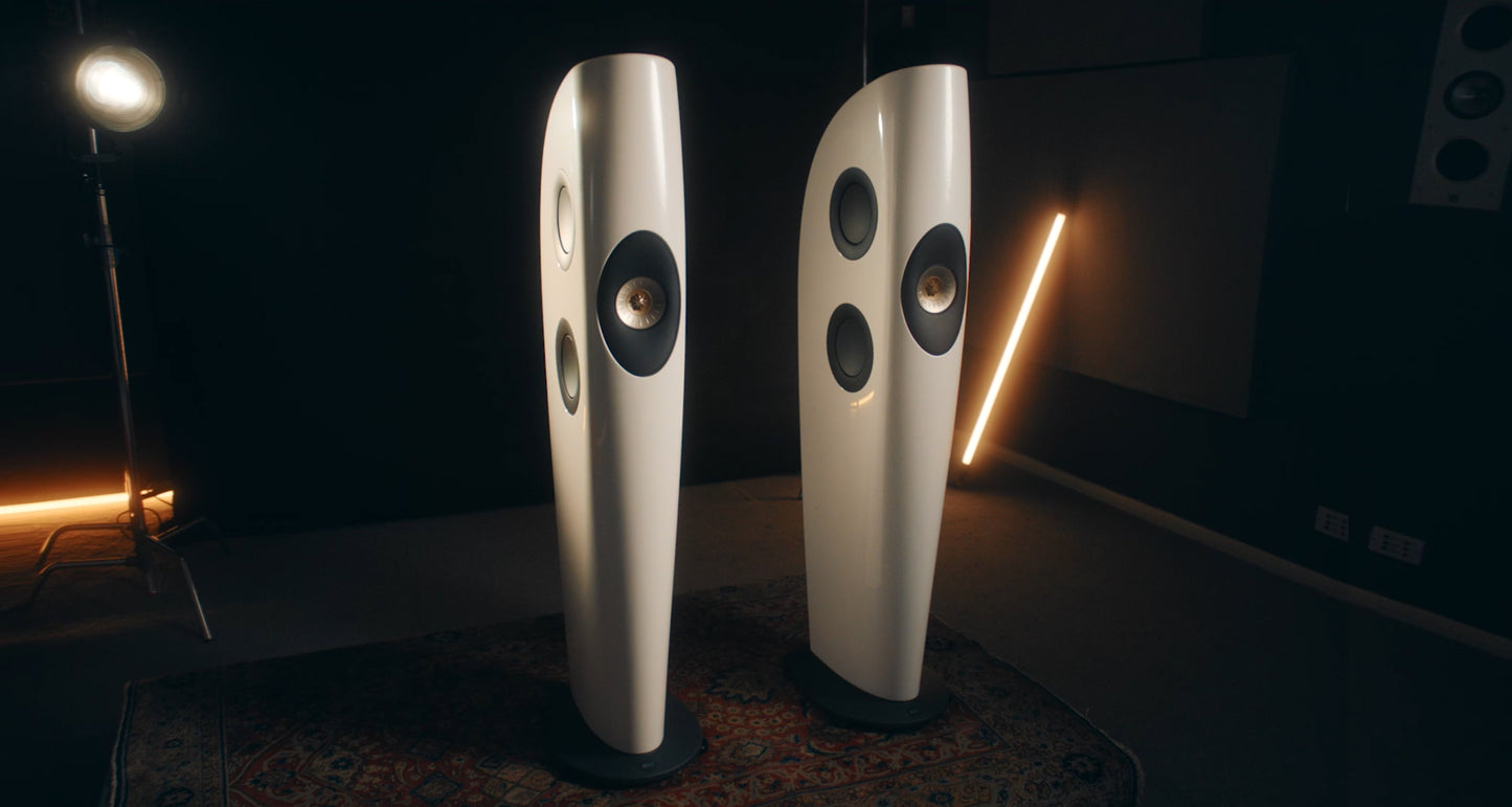 Pair of white KEF Blade Two Meta floor-standing speakers in a dark room with decorative lights.