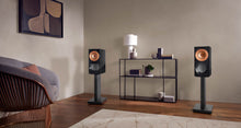 Two speakers on KEF Reference Reference 1 Stands in a living room setting with a shelf and chair.