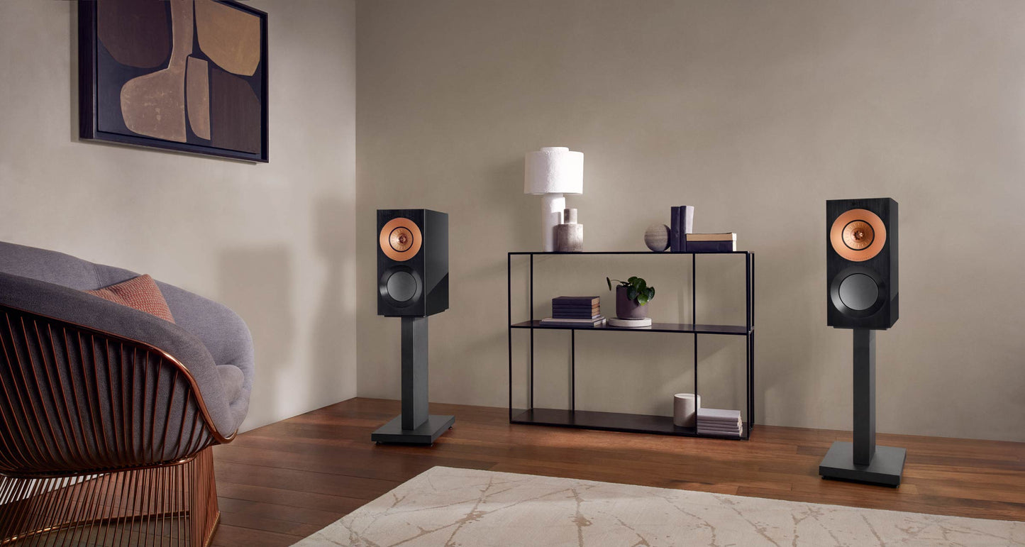 Two speakers on KEF Reference Reference 1 Stands in a living room setting with a shelf and chair.