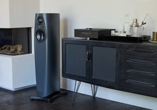 Tall charcoal Linn 343 floor-standing speaker with circular drivers beside a black media cabinet, part of a home audio.