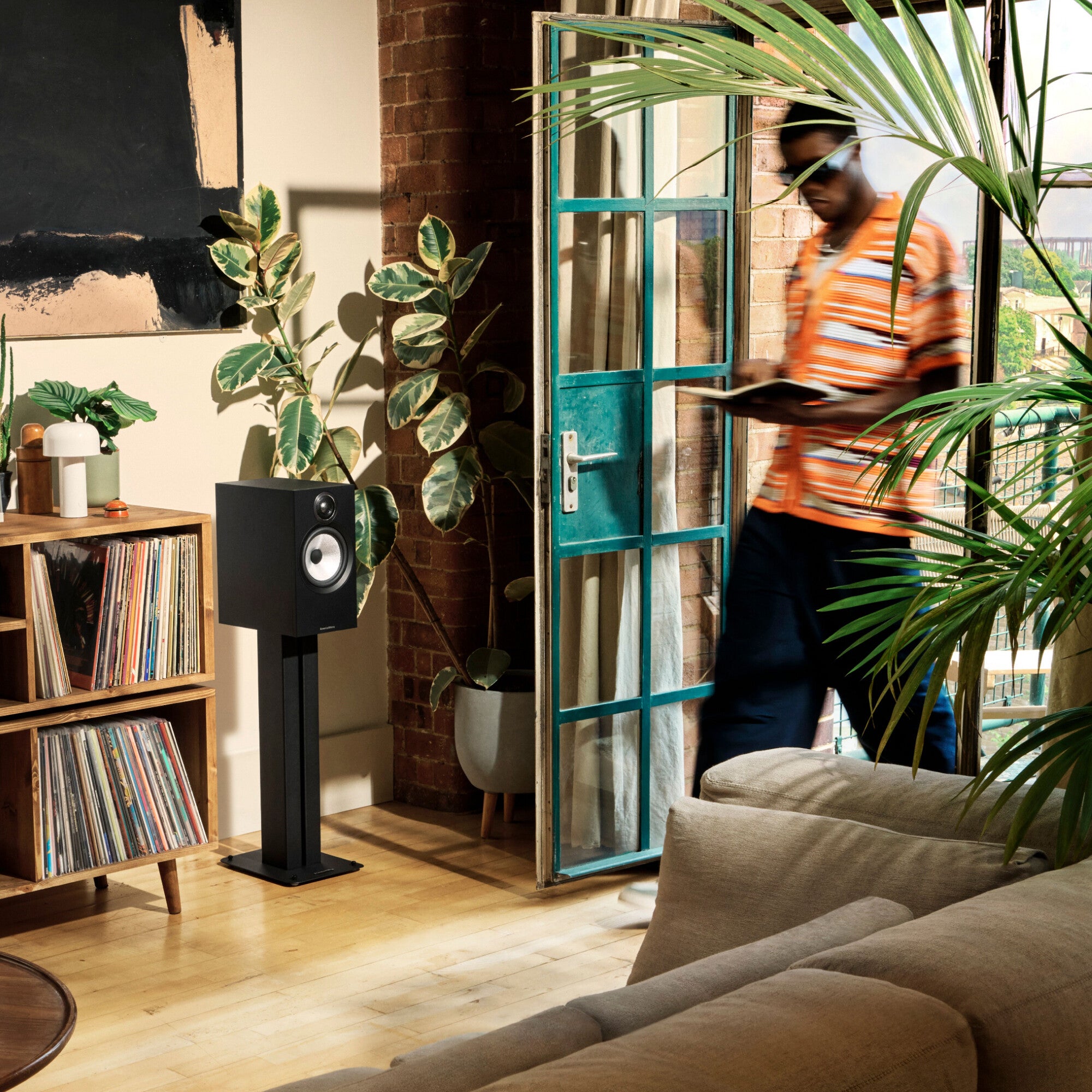 A person walking in a modern loft living room that has B&W 606 S3 Bookshelf Speaker in black finish standing next to a bookshelf.