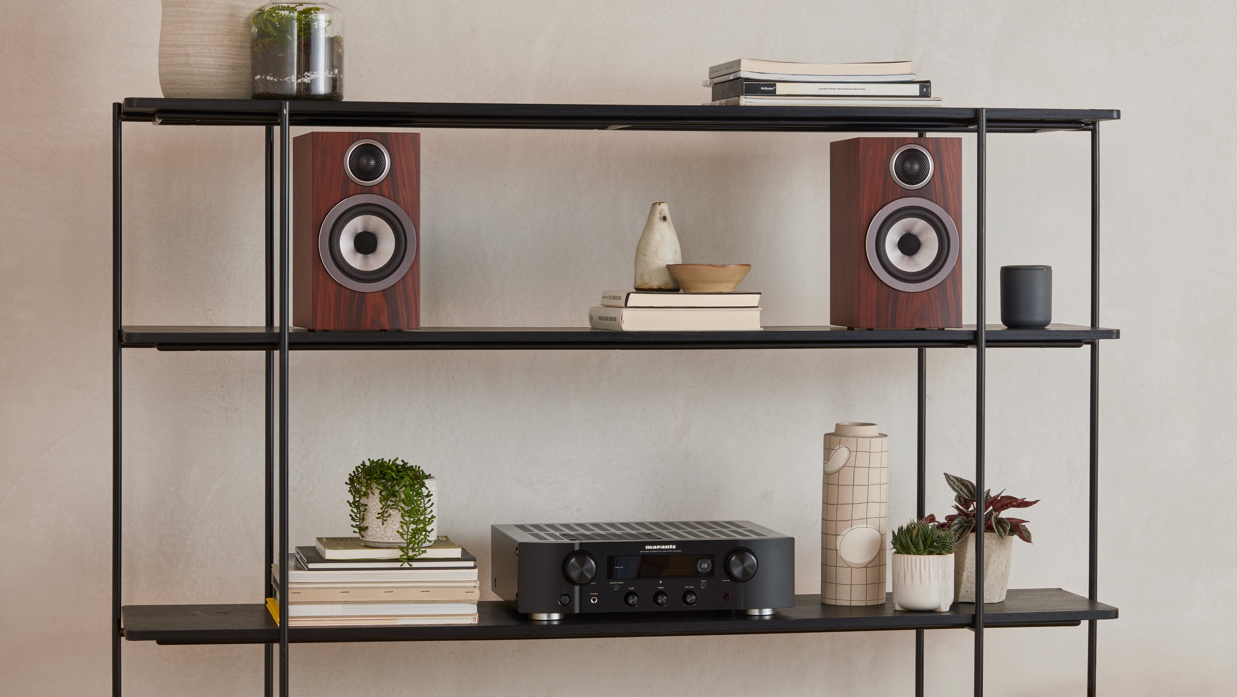 A pair of B&W 707 S3 Bookshelf Speakers in mocha finish placed on a modern open shelf in a living room.