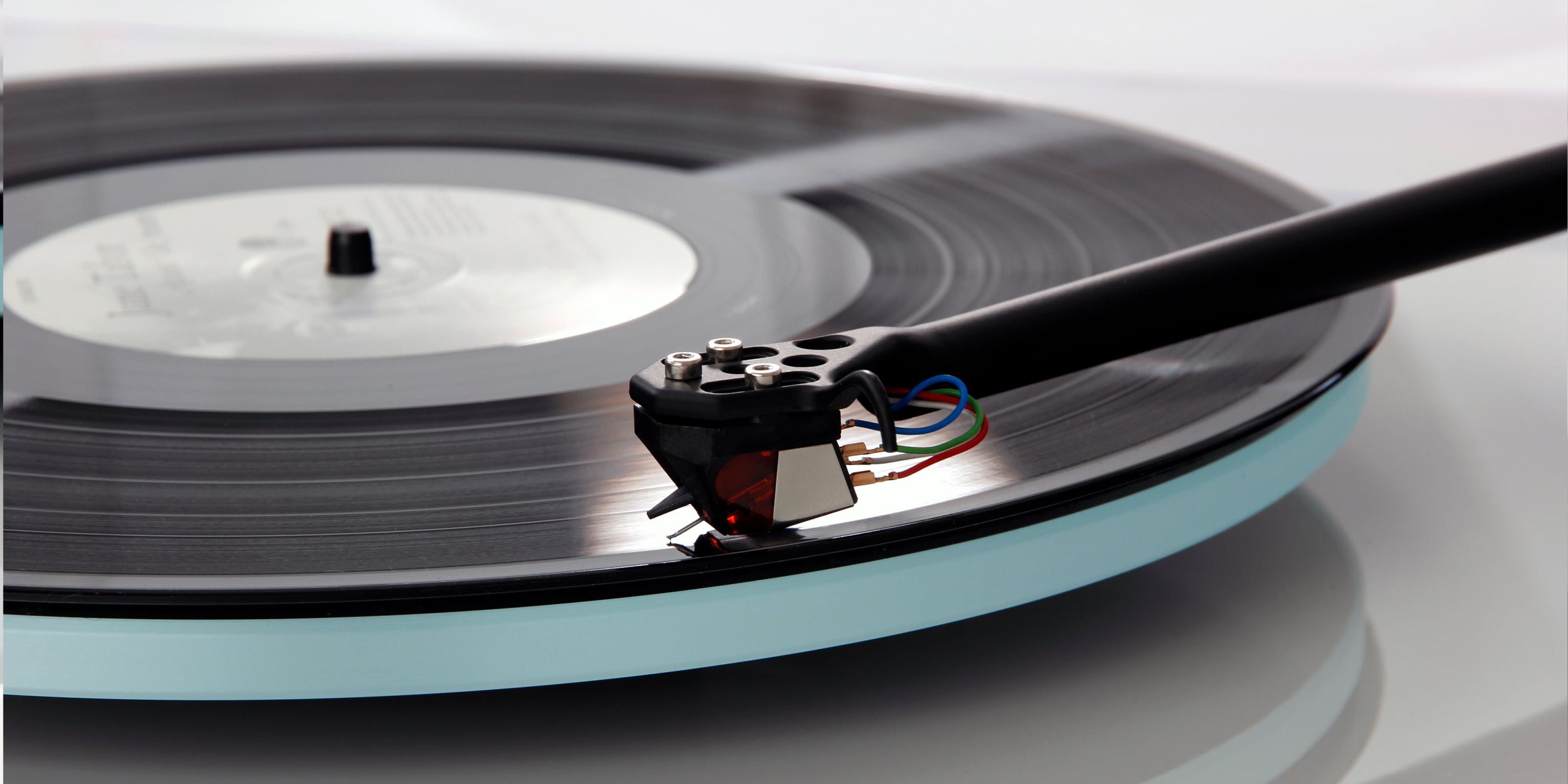 A close-up of a black vinyl record spinning on a Rega Planar 2 With Nd3 Cartridge, with the tonearm of a needle player resting across the top right.