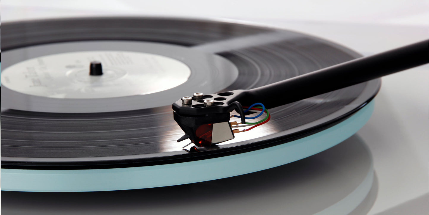 A close-up of a black vinyl record spinning on a Rega Planar 2 With Nd3 Cartridge, with the tonearm of a needle player resting across the top right.