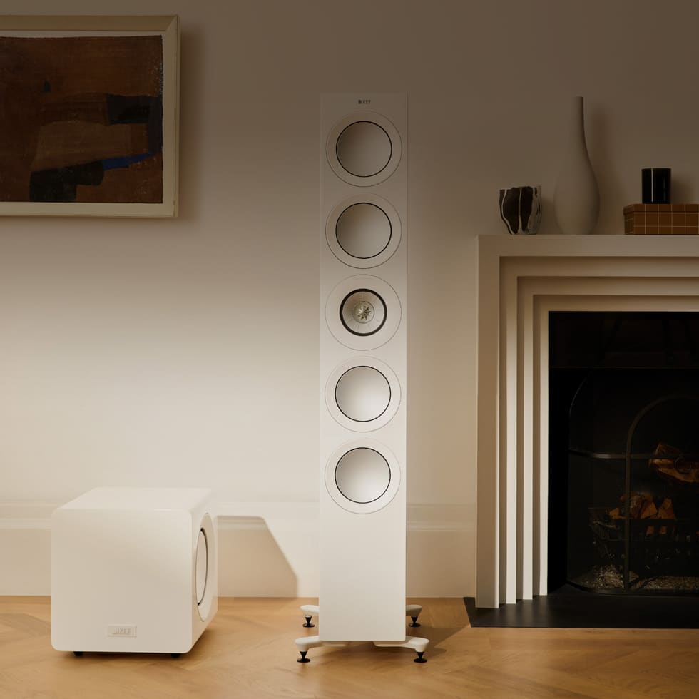 Tall white floor-standing speaker and a whilet KEF KC92 Dual 9” Subwoofer in a room with a fireplace and wooden floor.
