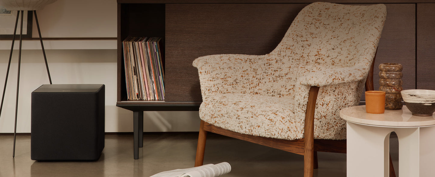 Patterned armchair in a room with a bookshelf, small table and a black KEF KUBE 10 MIE 10" Subwoofer.