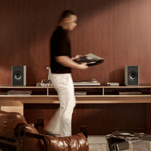 Person holding a record in a room with wooden walls and audio equipment including KEF Coda W Powered Speakers.