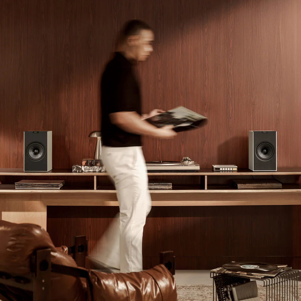 Person holding a record in a room with wooden walls and audio equipment including KEF Coda W Powered Speakers.