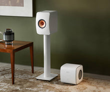 White speaker on a stand and KEF KC62 Subwoofer in a room with a green wall and wooden table.