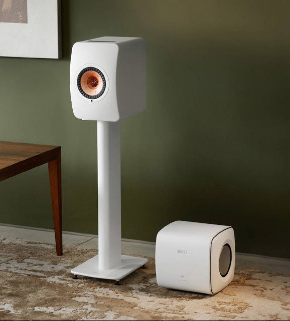 White speaker on a stand and subwoofer in a room with a green wall and wooden table.