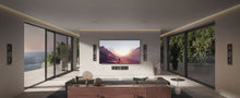 Modern living room with a large TV displaying a mountain scene with KEF Ci5160REFM-THX Reference In-Wall speakers installed, surrounded by large windows with a view of the ocean.