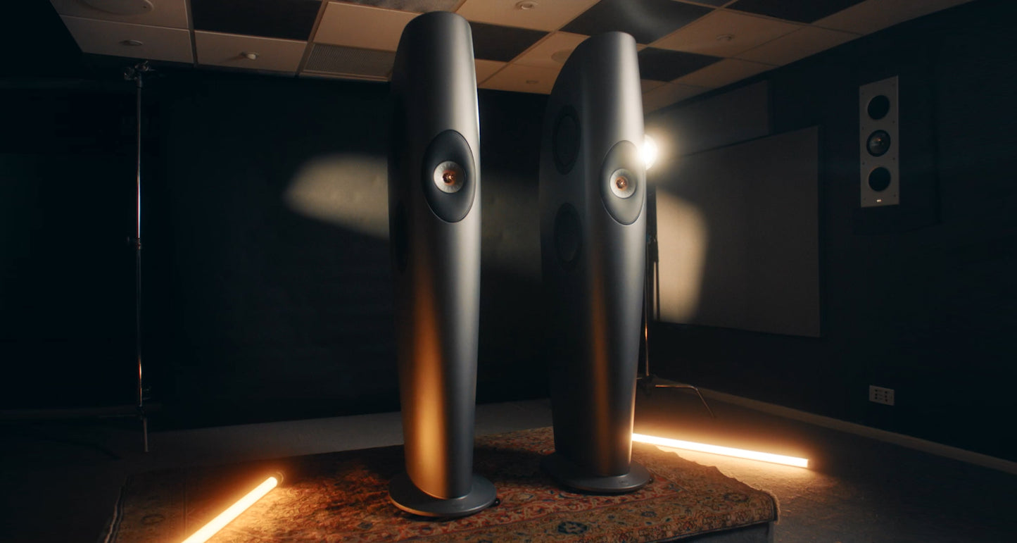 Two large KEF Blade One Meta speakers in a dark room with a projector screen.