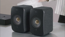 Two carbon black KEF LSX II Wireless Speakers on a surface with a blurred background