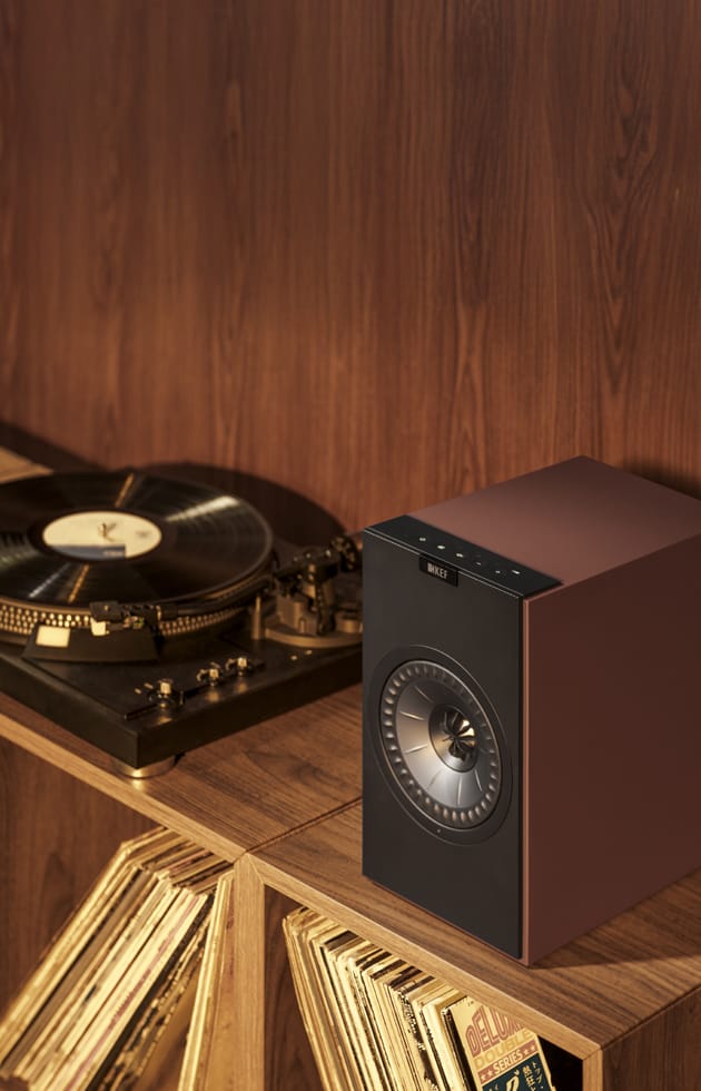Burgundy KEF Coda W Powered Speaker on a wooden surface with a vinyl record player and records in the background.