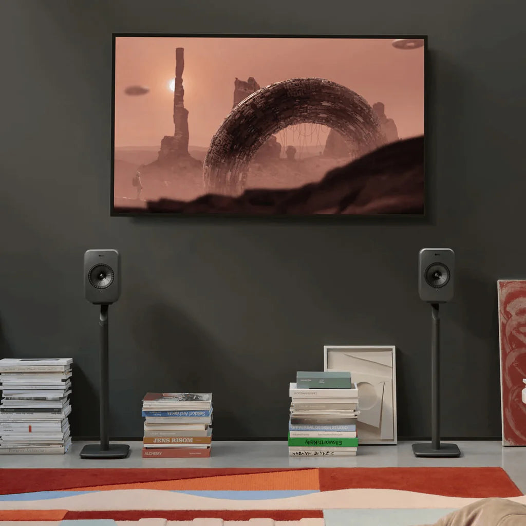 Modern living room with a TV displaying a desert scene, KEF speakers on KEF S1 Floor Stands for LSX II, and books on a shelf.