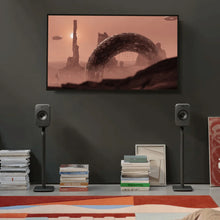 Modern living room with a TV displaying a desert scene, KEF speakers on KEF S1 Floor Stands for LSX II, and books on a shelf.