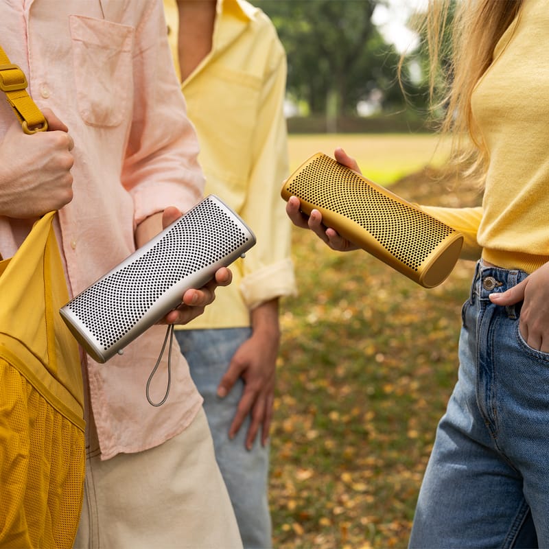 Two people holding KEF MUO Portable Bluetooth speakers outdoors with a natural background