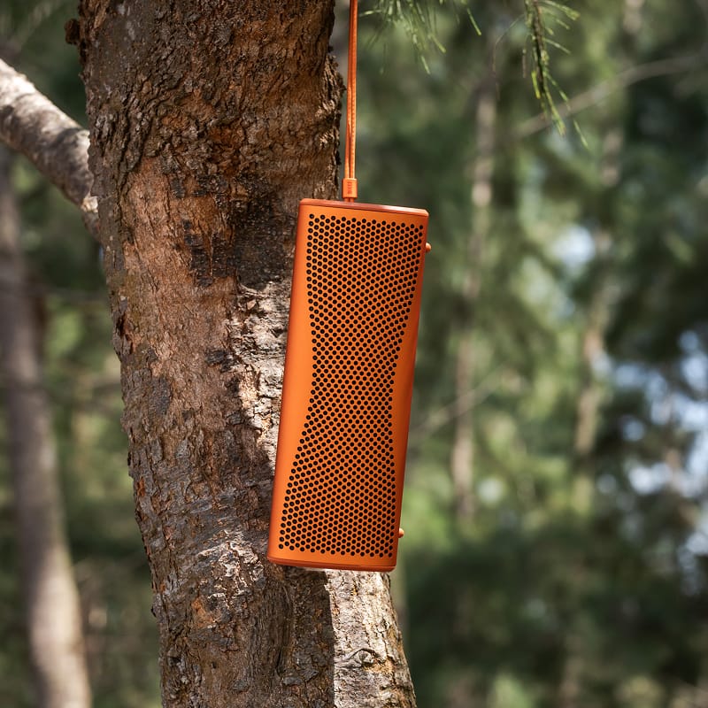 Orange rectangular KEF MUO Portable Bluetooth Speaker hanging from a tree branch with a blurred natural background
