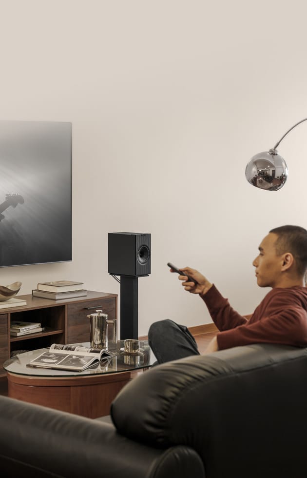 Man sitting on a couch in a living room with a TV and KEF Coda W Powered Speakers.